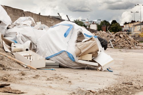 A large pile of discarded construction and building debris, including white plastic sheeting, cardboard boxes, and broken PVC pipes, is seen in an open, dusty outdoor area. The debris is haphazardly piled on the ground, with some materials partially unfolded or crumpled. In the background, there is a partially excavated earthbank and a distant view of an urban environment with several buildings, trees, and streetlights under an overcast sky with grey clouds. The scene suggests a site undergoing renovation or clearance, where rubbish removal services, such as those offered by House Clearance Hackbridge, might be required for proper disposal or cleaning of such waste materials.