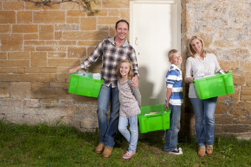A family of four stands outdoors in front of a stone wall, each holding a bright green plastic storage box used for rubbish collection or disposal. The father, dressed in a checked shirt and jeans, is smiling while holding one box in his right hand. Next to him, a young girl with long hair, wearing a grey patterned top and jeans, smiles and holds her own box, slightly leaning into him. To the right, a young boy with short blond hair, wearing a striped polo shirt and dark trousers, stands with one hand on his hip and the other holding a box. Beside him, a woman dressed in a white shirt and jeans, also holding a green box, looks at the camera with a neutral expression. The family appears to be preparing for rubbish removal, possibly as part of a house clearance service in Hackbridge, with the green boxes symbolising waste or recycling containers used in local rubbish disposal processes. The scene is illuminated by natural light, with a grassy patch in the foreground and the stone wall serving as a simple, neutral background.