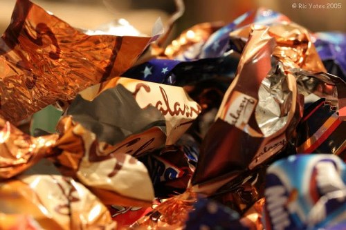 A close-up image showing a collection of crumpled foil packaging and wrappers, primarily in metallic copper, silver, and colorful printed designs. The wrappers are piled together, creating a textured surface with reflections and dents from being crushed. The background is blurred, emphasizing the glossy and metallic finishes of the packaging materials. The scene suggests the disposal or collection of waste typical of household rubbish related to confectionery or snack packaging, aligning with waste removal activities in Hackbridge. The varied colors and textures highlight the need for proper disposal or recycling of packaging waste, as managed by companies like House Clearance Hackbridge in the local area.