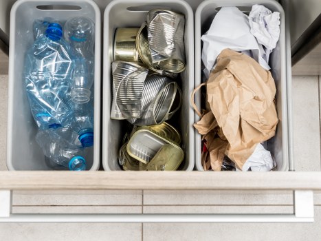 A three-compartment waste management bin situated on a tiled floor, with the left compartment containing clear plastic bottles, some with blue caps, the middle compartment filled with crushed or flattened aluminum cans in silver and gold tones, and the right compartment holding crumpled paper and brown paper bags, indicating sorting of recyclable rubbish. The bin appears to be part of a household waste collection setup, relevant to rubbish removal services in Hackbridge, with clean and organized separation of waste materials. The background includes a tiled surface, suggesting an indoor or garage environment commonly associated with waste disposal areas. The overall setup emphasizes proper waste sorting practices that a rubbish removal company like House Clearance Hackbridge might assist with, demonstrating standard recycling preparation during house clearance or rubbish collection processes.